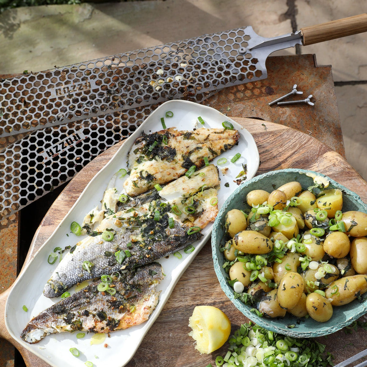 Grilled fish on a platter with a bowl of potatoes and green onions on a wooden table.