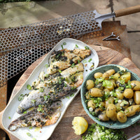 Grilled fish on a platter with a bowl of potatoes and green onions on a wooden table.