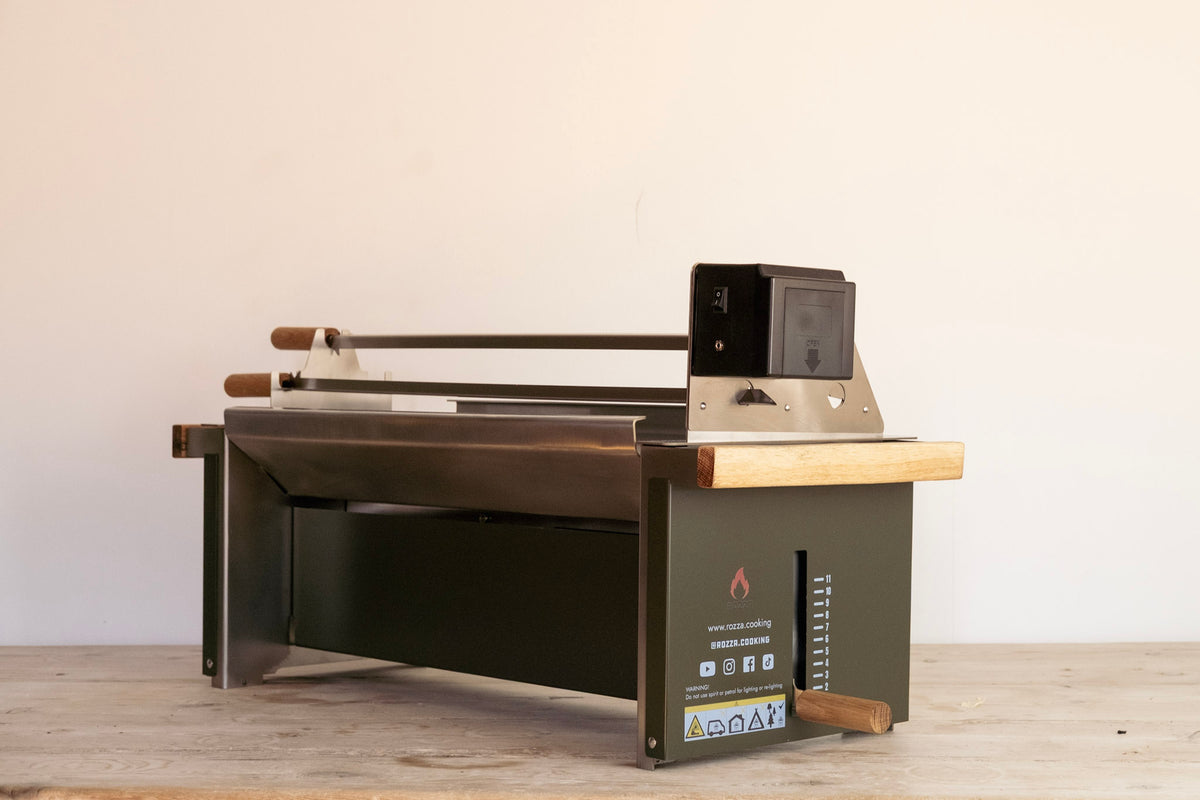 A compact outdoor propane grill with a dark green body, stainless steel grates, a light wood countertop, and a black control box on top, shown on a wooden surface against a plain beige wall.