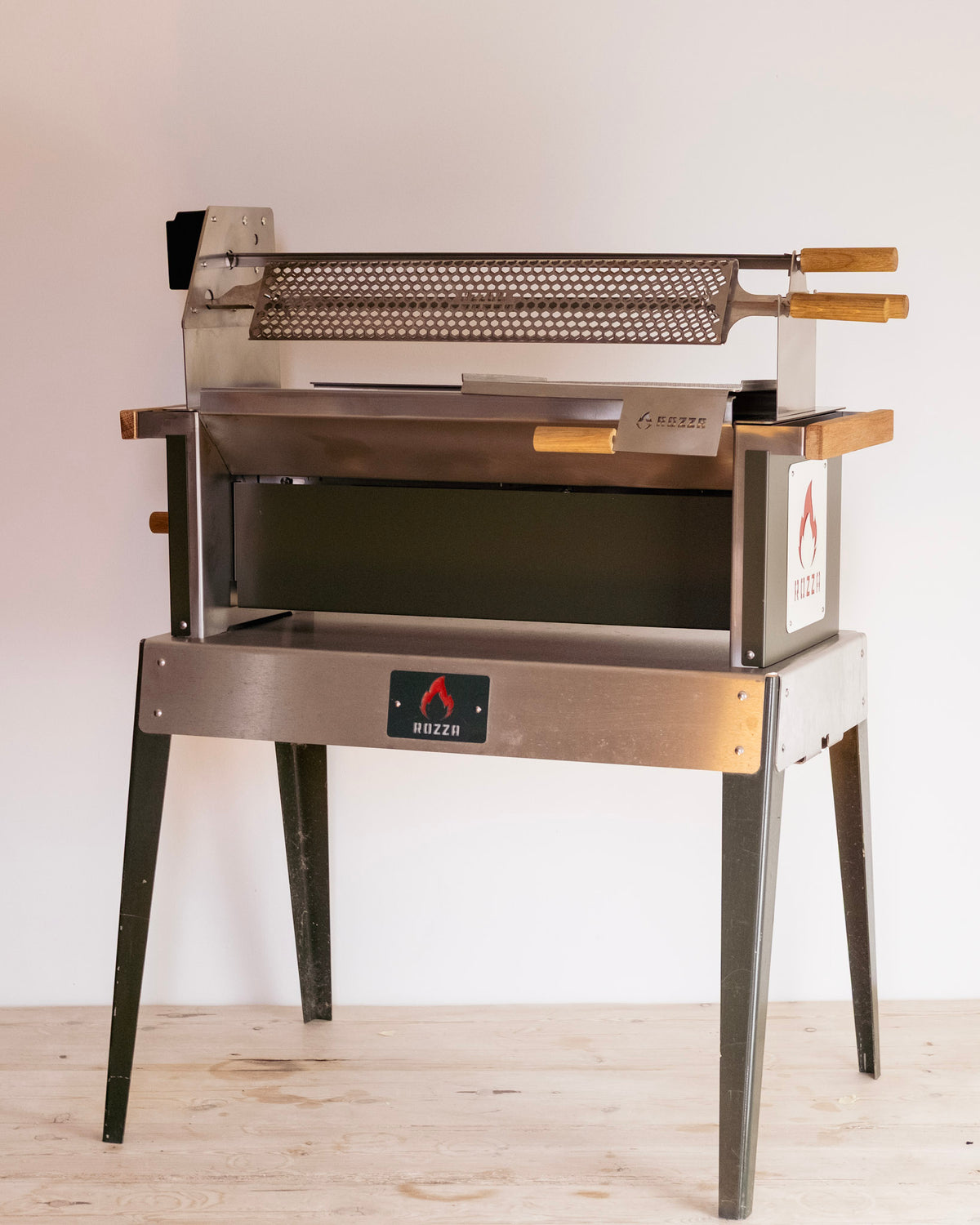 A commercial-style metal roasting or grilling stand with a perforated top rack, wooden handles, and four legs, photographed indoors against a plain light wall on a wood floor.