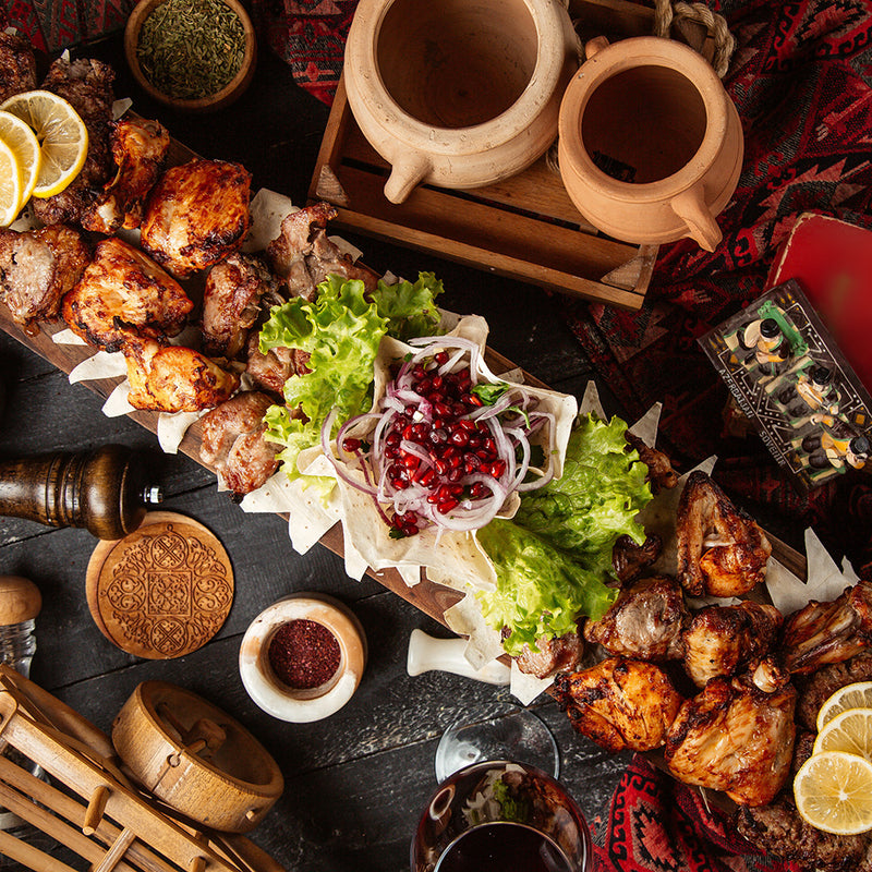 Plated dish of grilled meats with garnishes on a dark surface, surrounded by wooden containers and a patterned cloth.