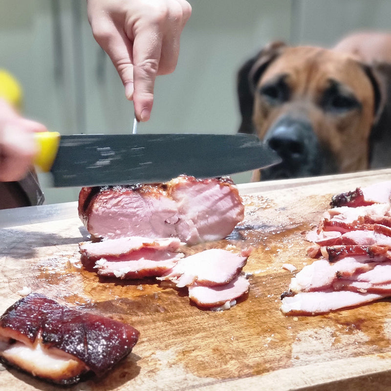 Person slicing ham on a wooden cutting board with a dog in the background.