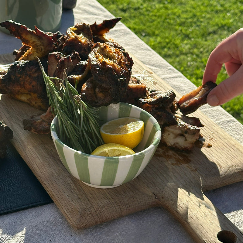 Wooden cutting board with roasted chicken, herbs, and lemons on a grassy outdoor setting.
