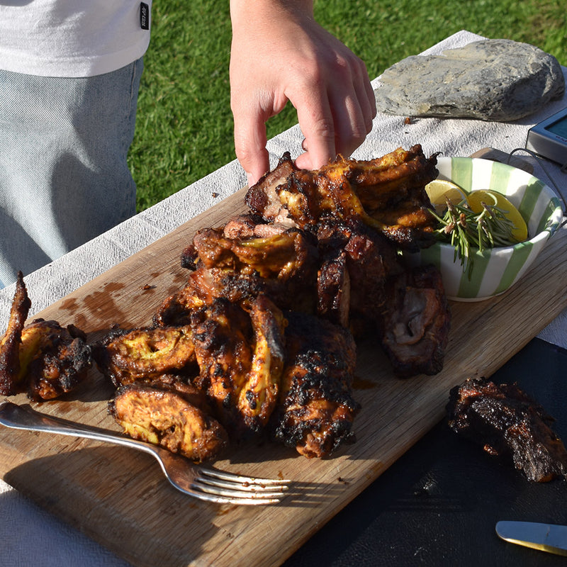 Grilled chicken wings on a wooden cutting board with a person's hand reaching for one, outdoors.