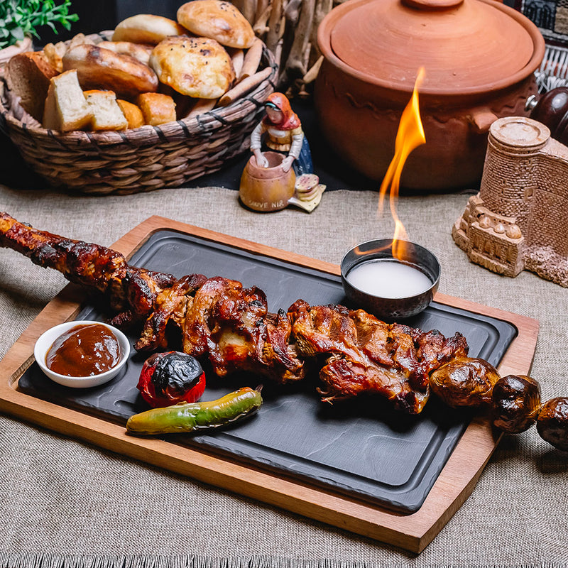 Grilled meat skewer on a tray with dipping sauces, surrounded by bread and a clay pot.